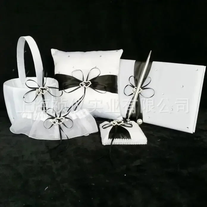 White wedding accessory set featuring a pillow, pen stand with pen, flower basket, and a guest book. Each item is adorned with black ribbons and decorative bows.