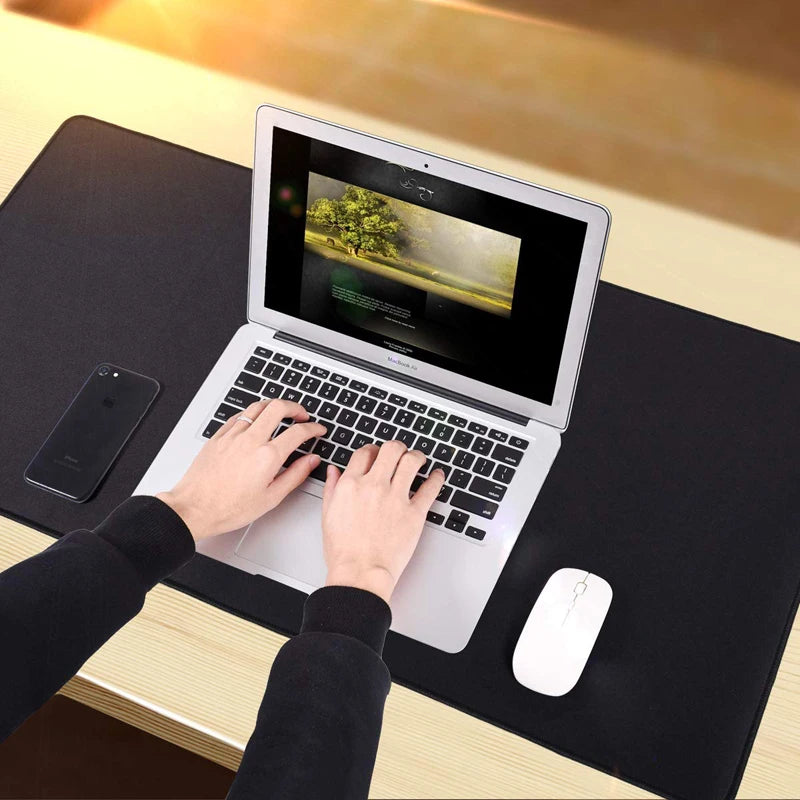 Hands typing on a laptop with a nature scene on the screen, placed on a black mat with a smartphone on the left and a white mouse on the right, in a softly lit setting.