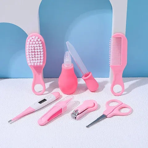 A set of pink baby grooming tools, including a soft brush, comb, digital thermometer, nasal aspirator, tweezers, nail clippers, and scissors, arranged on a white surface.