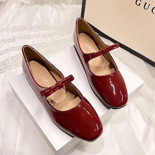 Elegant maroon patent leather Mary Jane flats with straps displayed on a white box. A shopping bag partially visible in the background on a white fabric surface.