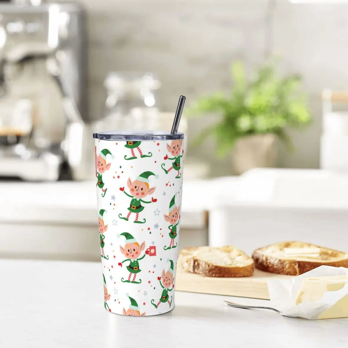 Festive tumbler with dancing elf patterns and a metal straw on a kitchen counter. Nearby, a cutting board holds partially buttered toast. Blurred kitchen items in the background.
