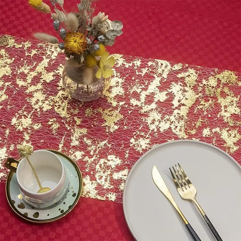 A decorative table setting with a gold-patterned red tablecloth, a vase of dried flowers, a teacup with saucer and spoon, and a gray plate with gold cutlery.
