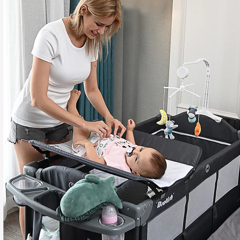A woman dressed in a white t-shirt and gray shorts is changing a baby’s diaper on a changing station attached to a playpen. A mobile with toys hangs above the baby.