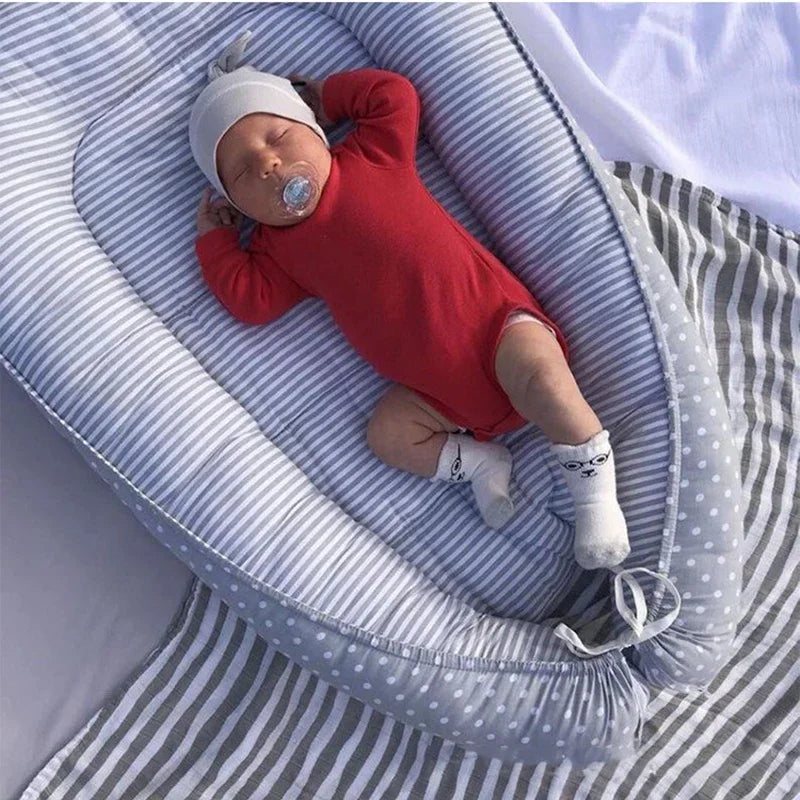Sleeping baby in a red onesie and gray hat, with a pacifier, lies in a gray and white striped baby nest. The nest is placed on a matching gray-striped and polka dot blanket.