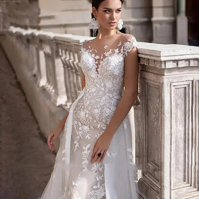 Woman in a detailed floral lace wedding dress stands against a stone railing, looking to the side. The dress features an illusion neckline and sheer overlay with intricate patterns.