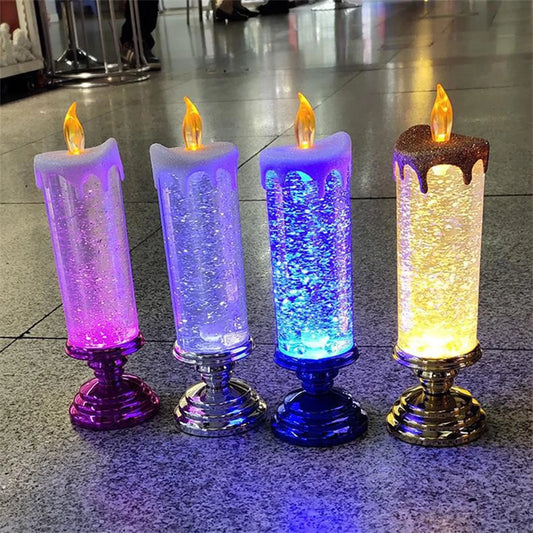 Four decorative LED candles with glittering liquid interiors and glowing flames, in pink, silver, blue, and yellow, stand on shiny bases on a reflective surface.