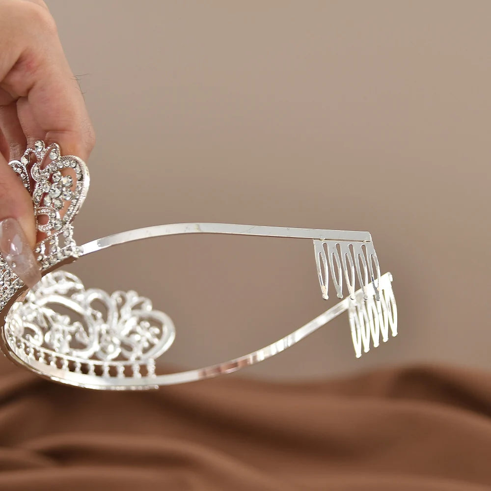 A hand holds a silver tiara adorned with intricate patterns and embedded with small sparkling gems, set against a soft beige background.