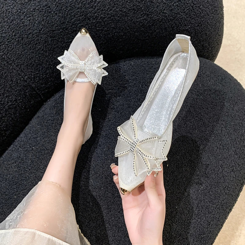 Transparent shoes adorned with rhinestone bows and gold-capped pointed toes, resting on a textured black surface. One is worn on a foot, the other held in hand.
