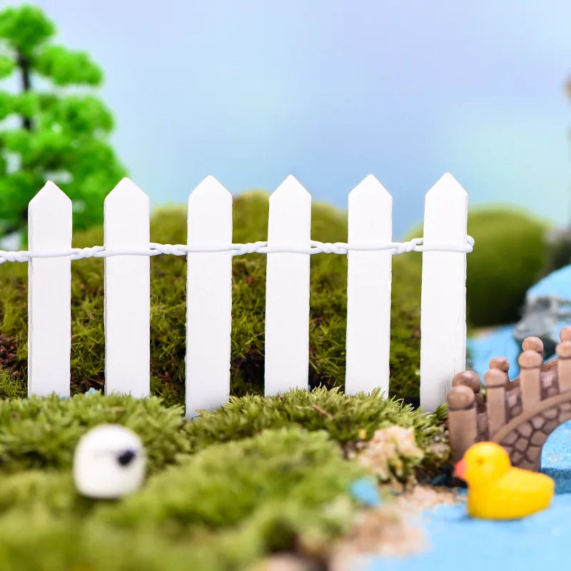 Miniature garden scene with a white picket fence, green moss ground cover, a small bridge, and a yellow duck figurine near a water feature, against a clear blue sky.