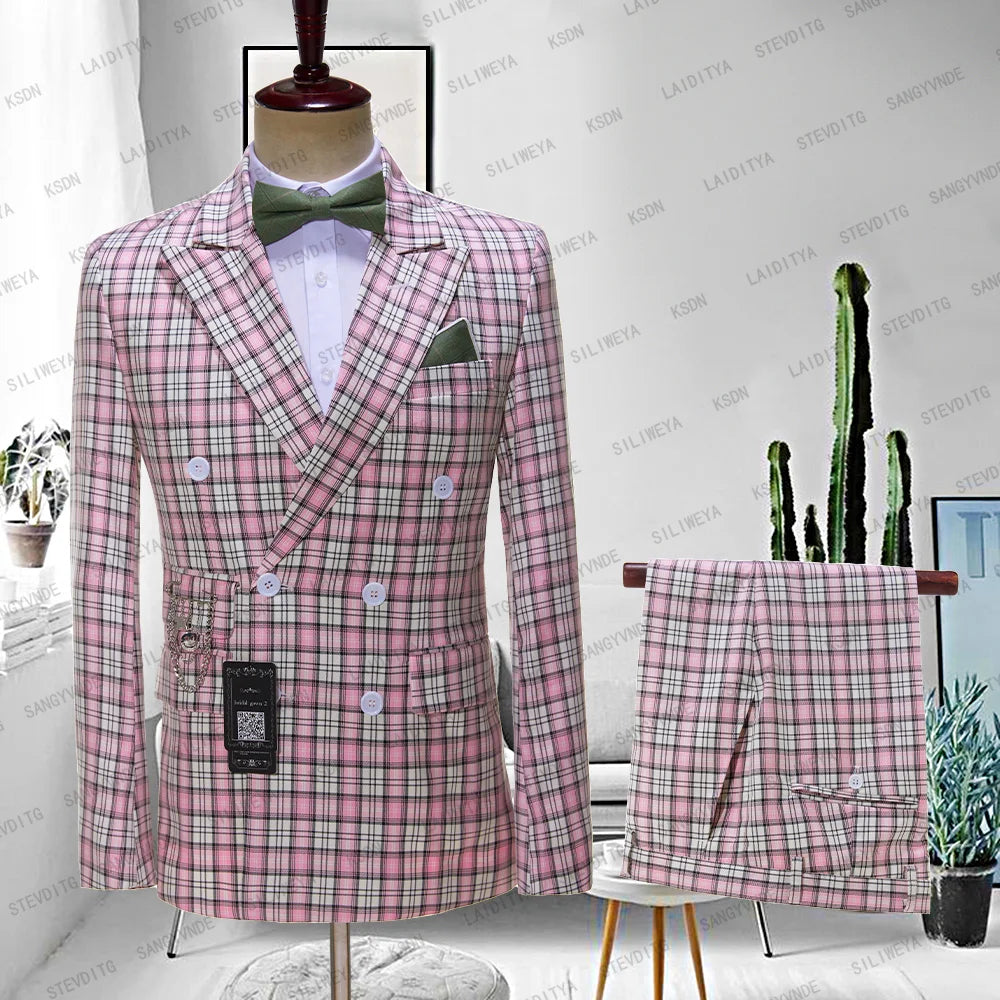 Pink plaid double-breasted suit with a green bow tie and pocket square on a mannequin. Matching pants draped nearby. Room includes a plant and abstract wall art.
