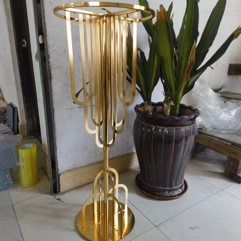 Golden art deco-inspired metal plant stand with elongated curves, positioned on a tiled floor next to a tall potted plant in a dark brown pot near a wall.
