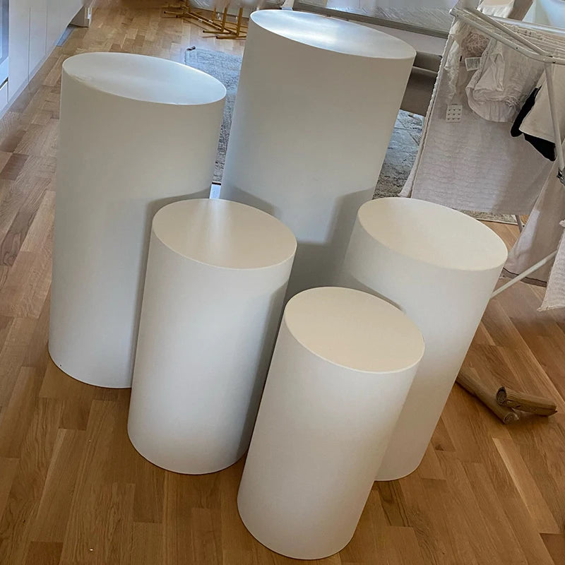 Five white cylindrical pedestals of varying heights are arranged on a wooden floor in a room. Draped fabric and furnishings are visible in the background.