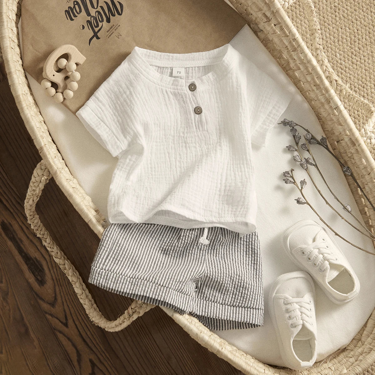 Baby outfit includes a white shirt, striped shorts, and white shoes in a woven basket. A wooden toy and dried flowers are also present on a wooden surface.