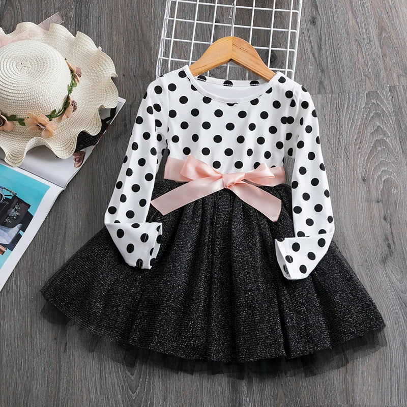 A polka-dot dress with a black skirt and pink bow at the waist, hung on a wooden hanger. A straw hat and a magazine are laid nearby on a wooden floor.