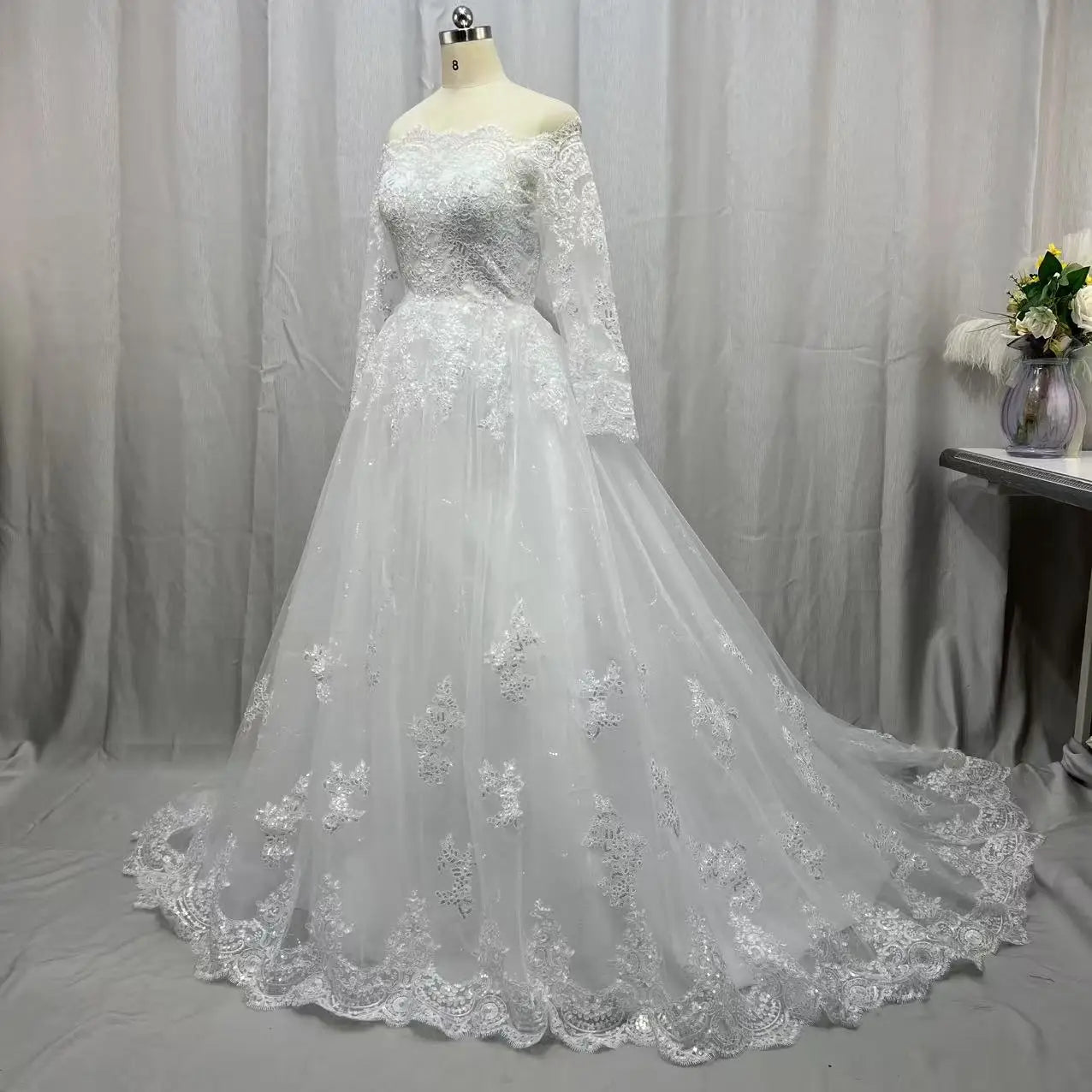 Elegant off-shoulder wedding dress on a mannequin, featuring intricate lace patterns, long sleeves, and a flowing train. It stands against a soft draped background with a flower vase nearby.