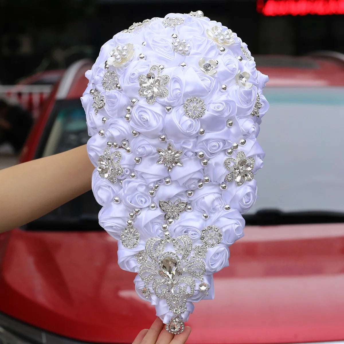 Hand holding a teardrop-shaped bridal bouquet with white satin roses and silver jeweled accents, in front of a red car.
