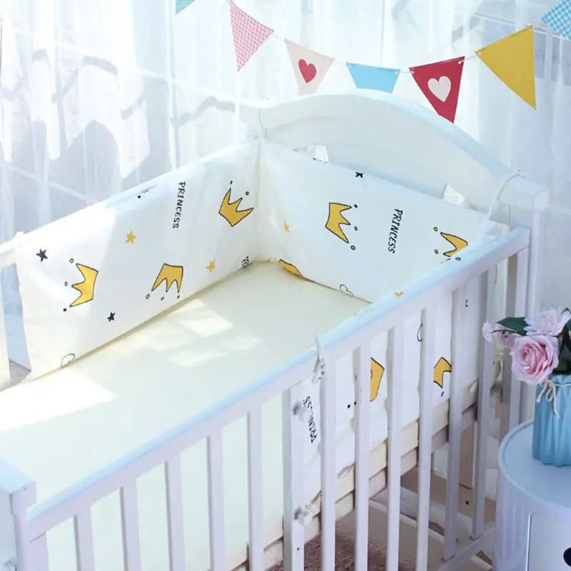 A white baby crib with crown-patterned cushions labeled "Princess," decorated with colorful bunting. A blue vase with pink flowers is on a table beside it.