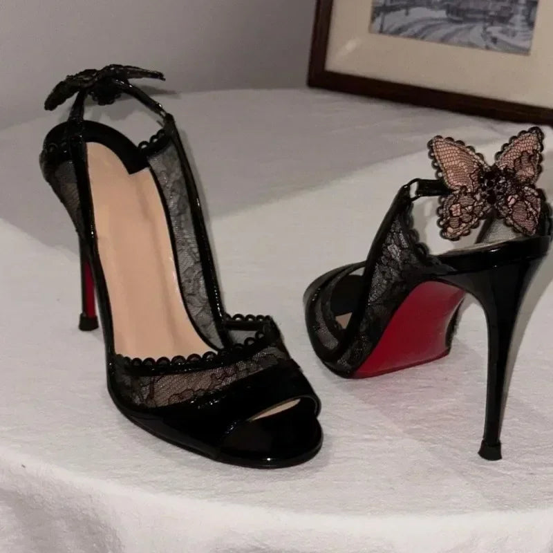 Elegant black high heels with lace detailing and butterfly accents on the back. The shoes feature a red sole and are displayed on a white table with a framed picture in the background.