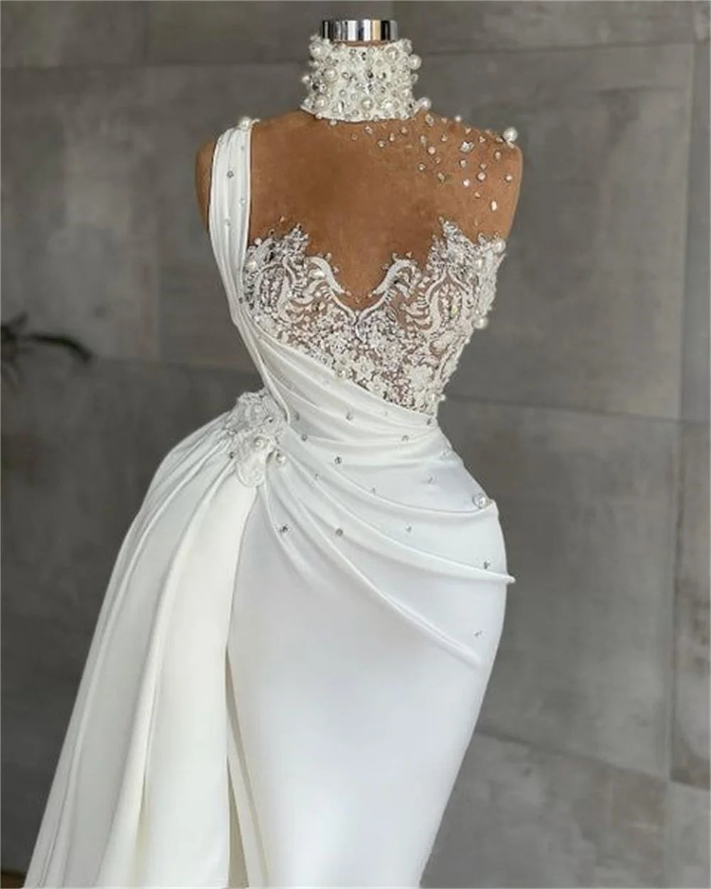 Elegant white gown on a mannequin, featuring intricate pearl and lace detailing on the bodice, an asymmetric strap, and a high neck adorned with pearls, against a gray background.