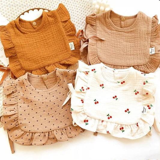 Four ruffled baby bibs are displayed. Two are solid colors—rust orange and beige—while the others feature patterns: one with black polka dots and the other with small red cherries.
