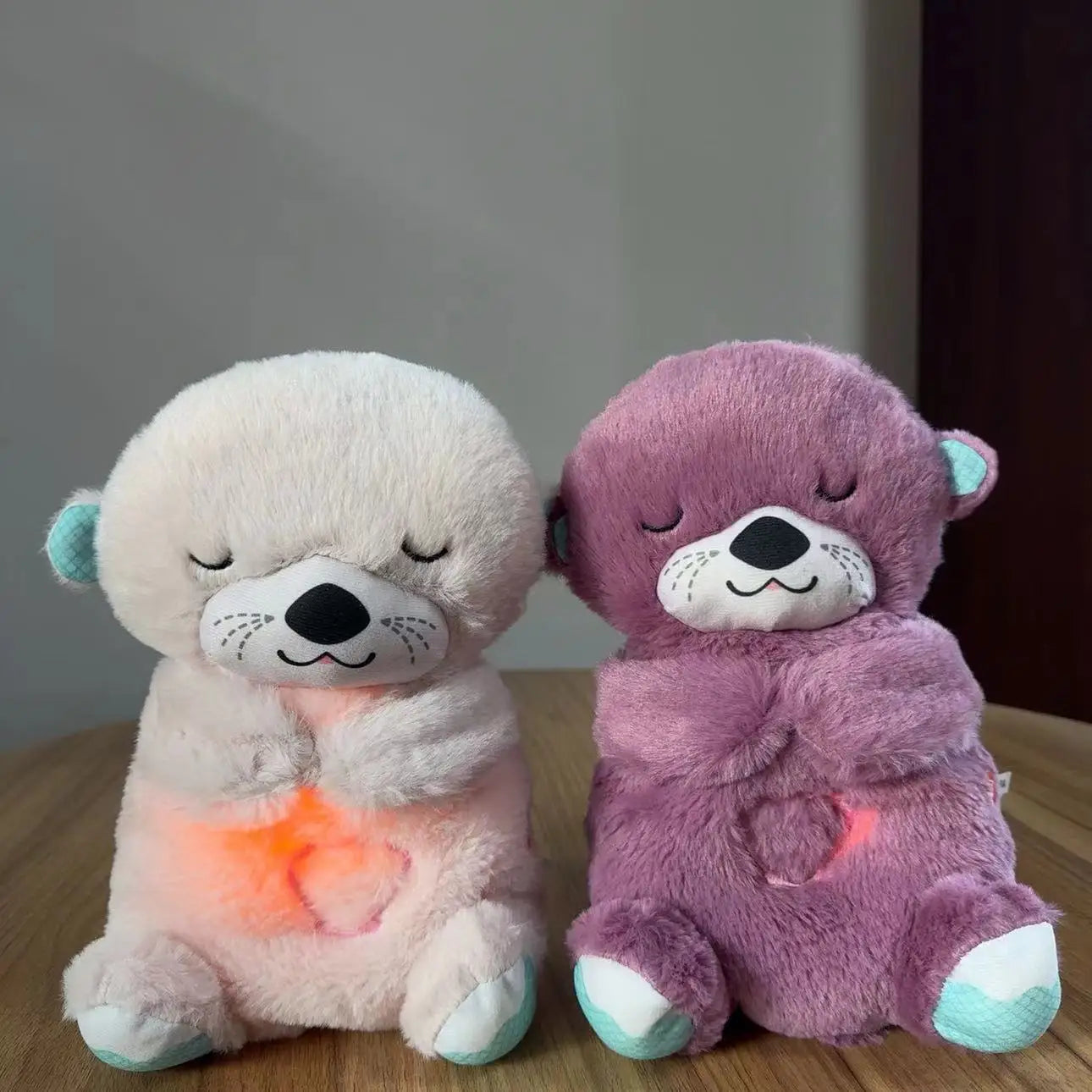 Two plush toys, one white and the other purple, sit on a wooden table. Both have closed eyes with a gentle smile, holding a glowing heart on their chests.