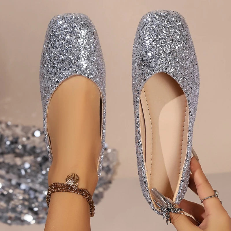 Silver glittery flats with a rounded toe design shown both on a foot and held by a hand. The person wears an anklet with a shell charm. Background is softly out of focus.