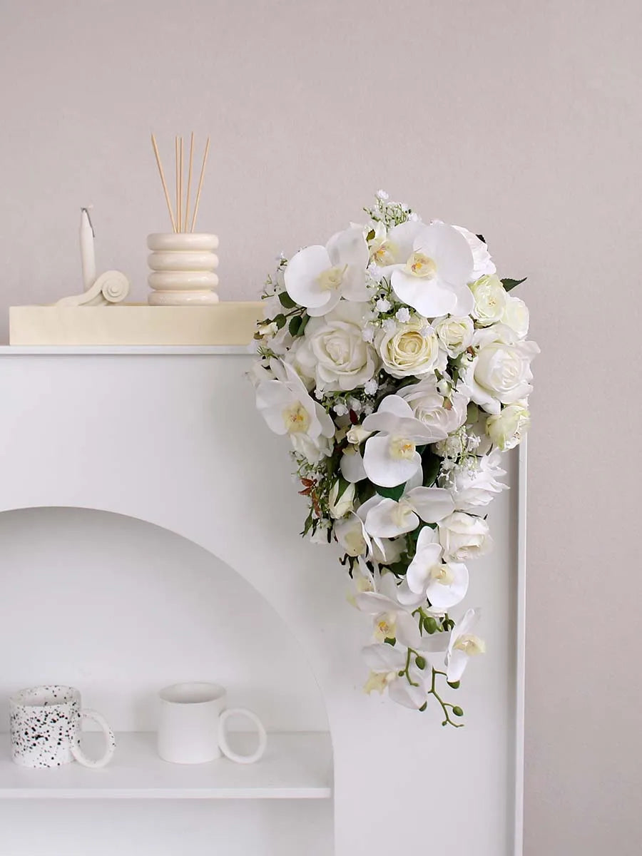 A cascading bouquet of white orchids and roses hangs over a white mantel, accompanied by a decorative candle, reed diffuser, and two mugs on a shelf below.