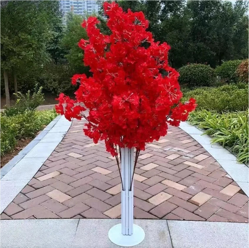 A vibrant red maple tree in a sleek white planter stands on a patterned brick pathway, surrounded by lush greenery and trees, with an urban skyline visible in the background.