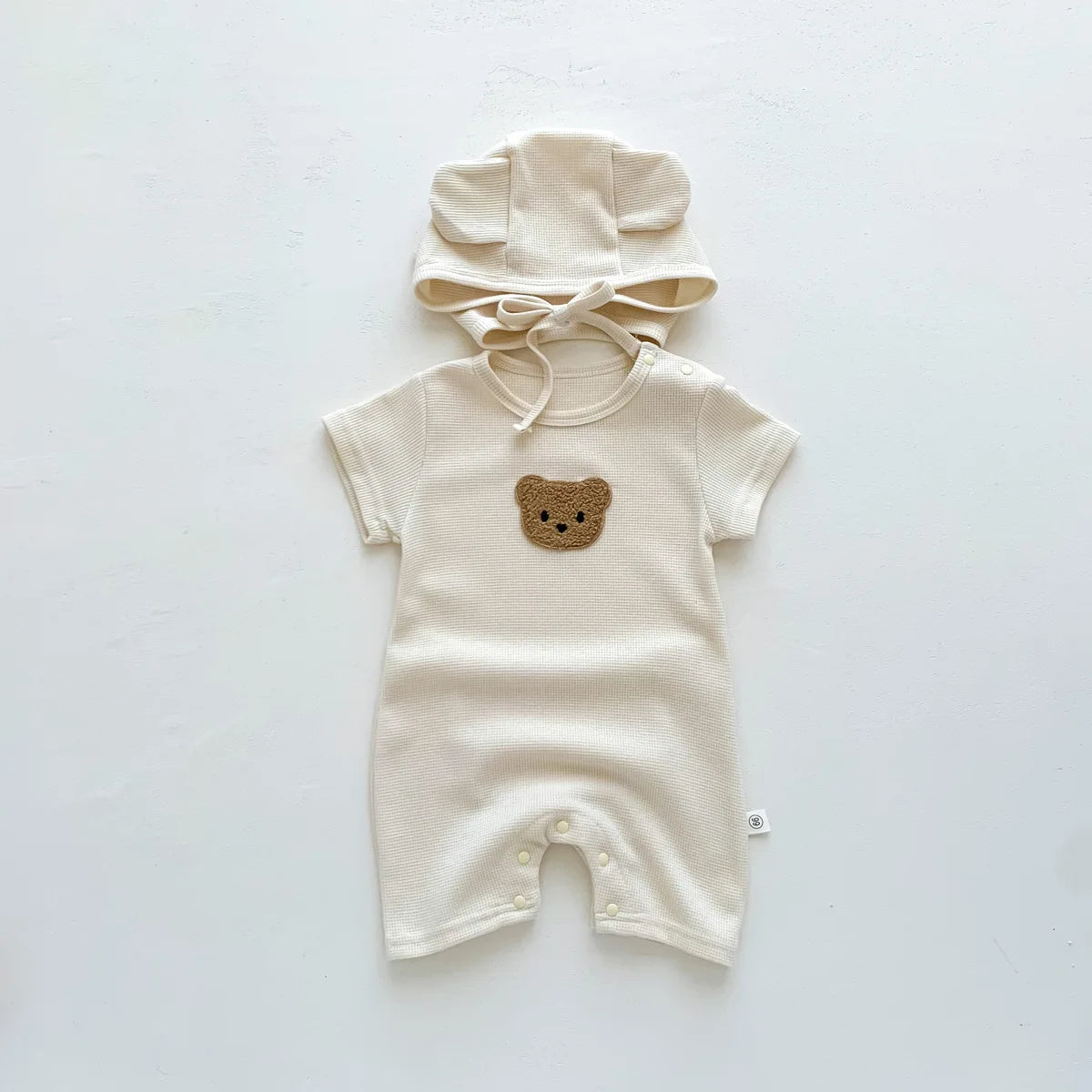 Beige baby romper with short sleeves, featuring a brown embroidered bear face on the chest. Comes with a matching bonnet with bear ears and a tied chin strap.