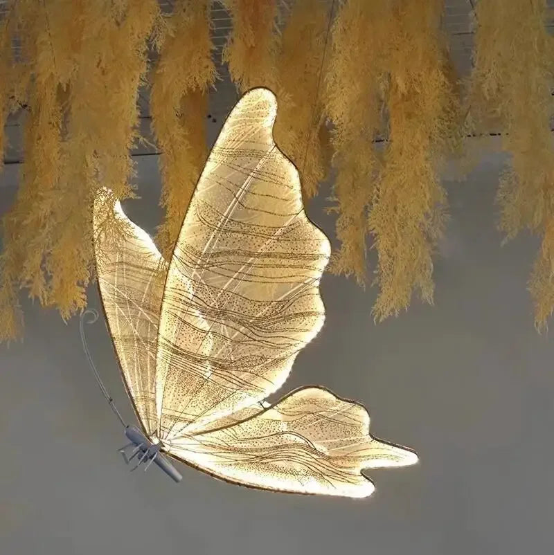 A glowing butterfly-shaped light fixture with delicate, translucent wings hangs beneath golden, fern-like foliage against a soft gray background.