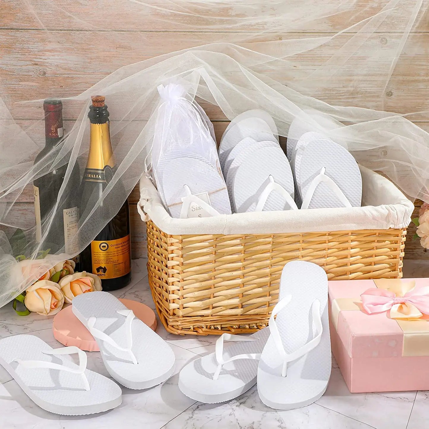 A wicker basket of white flip-flops, surrounded by wine bottles, roses, and a pink gift box with a bow, set on a marble surface with sheer fabric draped above.