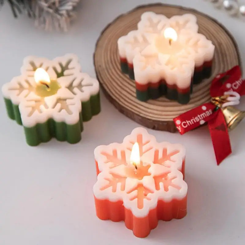 Three lit snowflake-shaped candles in Christmas colors sit on a white surface and wooden coaster, adorned with a small bell and red ribbon, creating a festive ambiance.