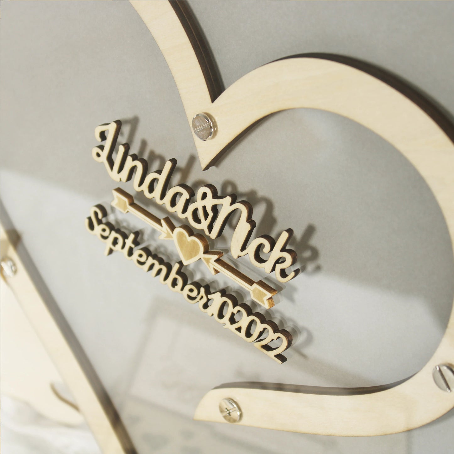 Wooden heart frame with carved text "Linda & Nick September 2022" and small heart detail, attached with screws on a gray background.