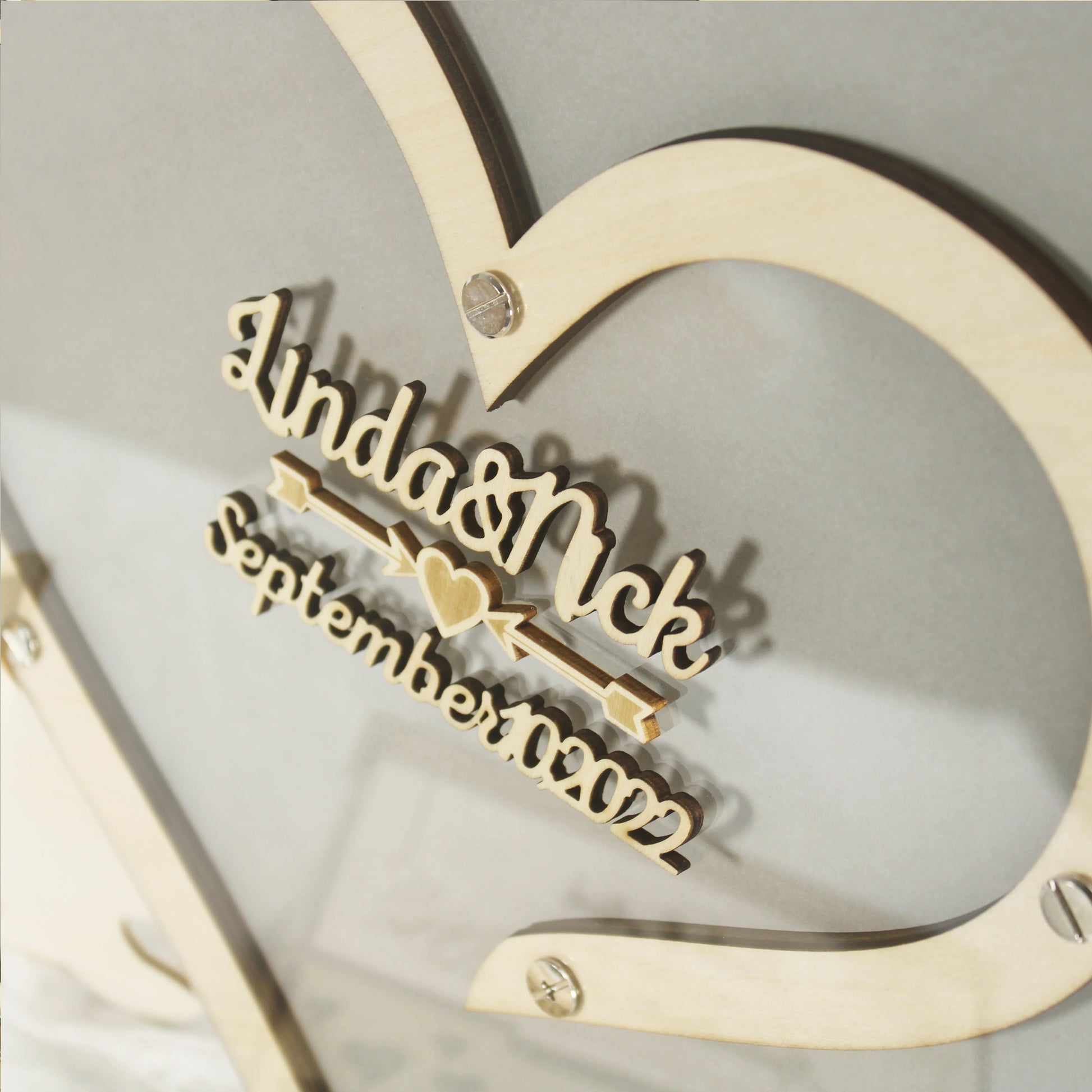 Wooden heart frame with carved text "Linda & Nick September 2022" and small heart detail, attached with screws on a gray background.