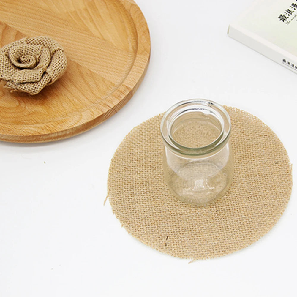 A small glass jar sits on a round burlap mat. Nearby, a wooden tray holds a burlap rose, and an open book is partially visible on a white surface.