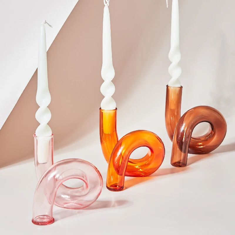 Curved glass candlesticks in shades of pink and orange, holding white twisted candles, are arranged on a light surface, creating a modern, artistic display.