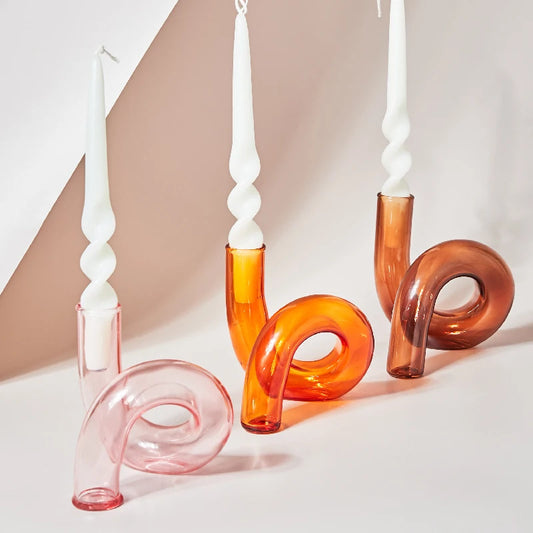 Curved glass candlesticks in shades of pink and orange, holding white twisted candles, are arranged on a light surface, creating a modern, artistic display.