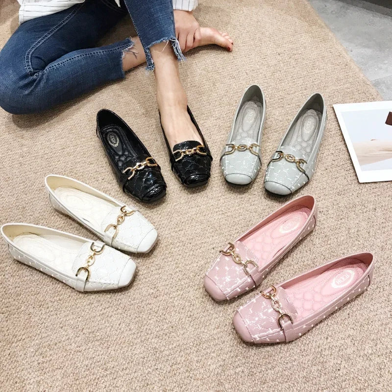 Person wearing black loafer with gold chain, surrounded by five other colorful loafers on carpet. Jeans-clad legs visible, creating a casual and stylish display.