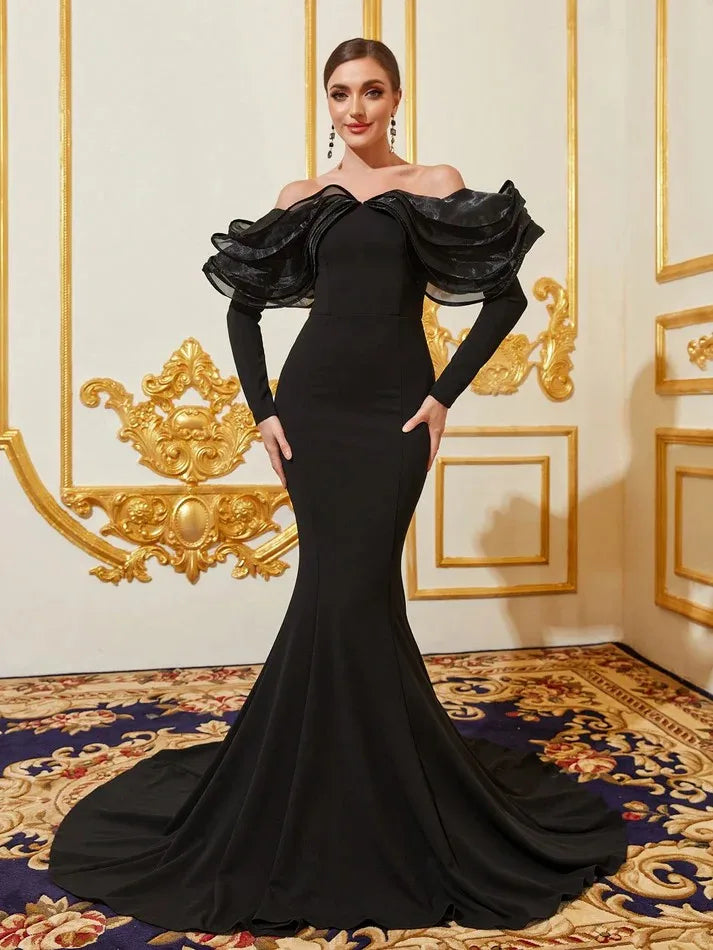 Elegant woman in a black off-the-shoulder gown with ruffled sleeves stands in an ornate room with gold details and a patterned carpet.