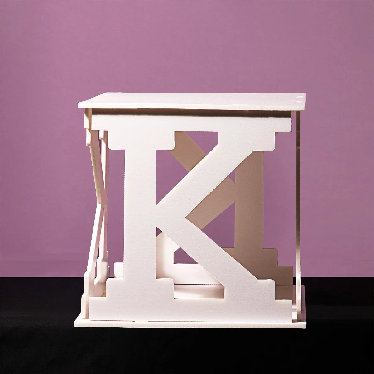 White stool shaped like the letter "K" against a purple background, with a minimalist design and resting on a black surface.