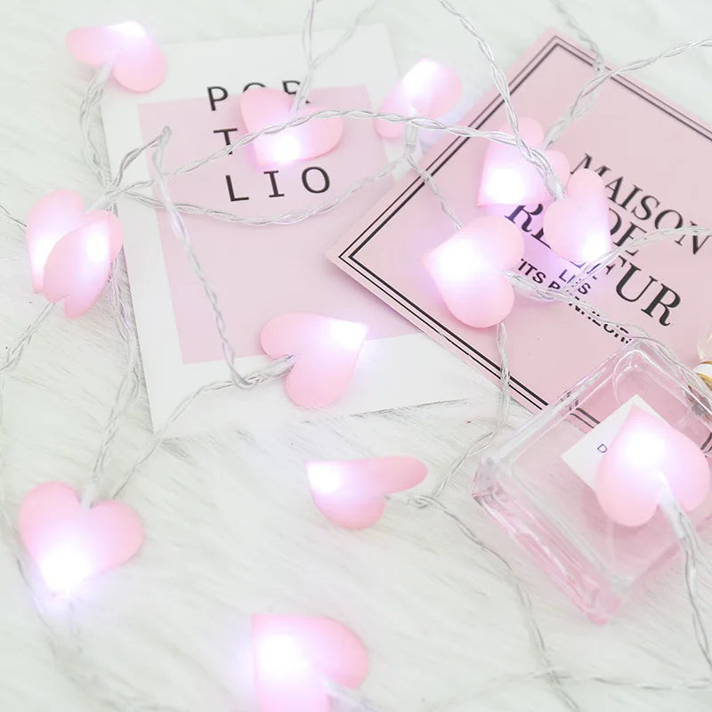 Pink heart-shaped fairy lights on a white surface, intertwined with pink postcards and a clear perfume bottle. The lights create a soft, romantic glow.