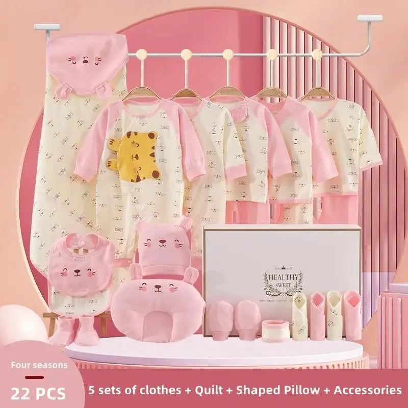 Set of baby clothes and accessories in soft pink and cream hues. Includes five outfits, quilt, shaped pillow, bib, booties, and more, displayed on hangers and a table.