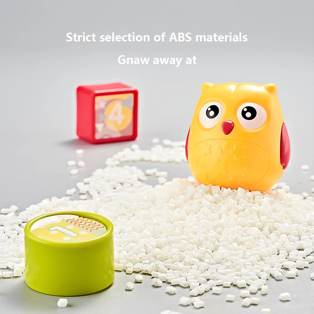 A yellow owl toy and green cap sit on white ABS plastic pellets. A red cube with the number 4 is in the background. Text above reads, "Strict selection of ABS materials. Gnaw away at."