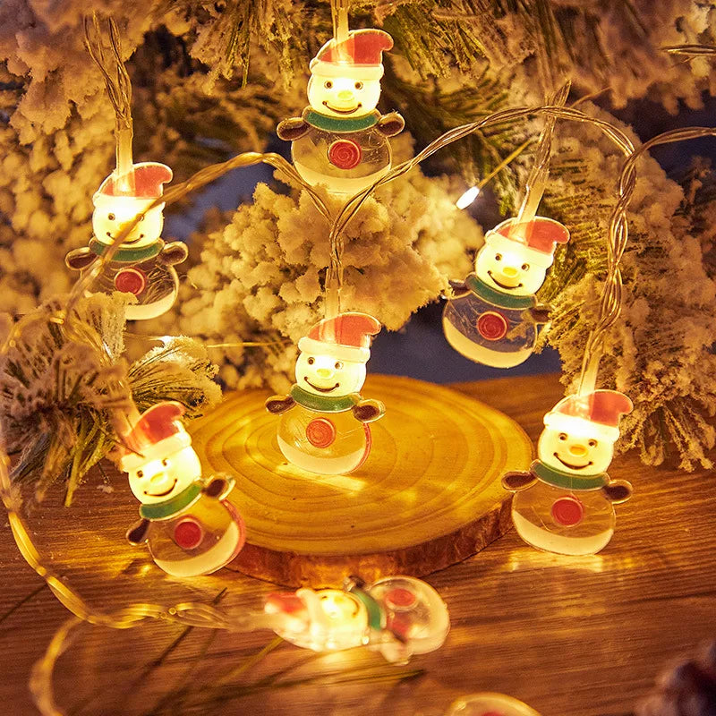 Glowing snowman string lights with red hats and green scarves adorn a snow-covered pine branch, casting a warm, festive ambiance on a wooden surface.