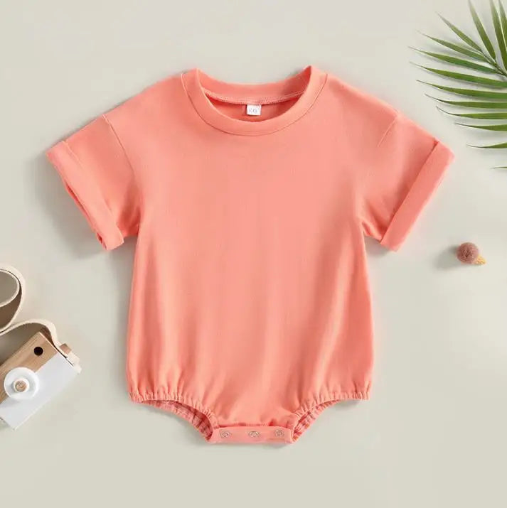 Peach-colored baby romper with short sleeves and snap buttons at the bottom, surrounded by a small toy camera and a sprig of green leaves on a light background.