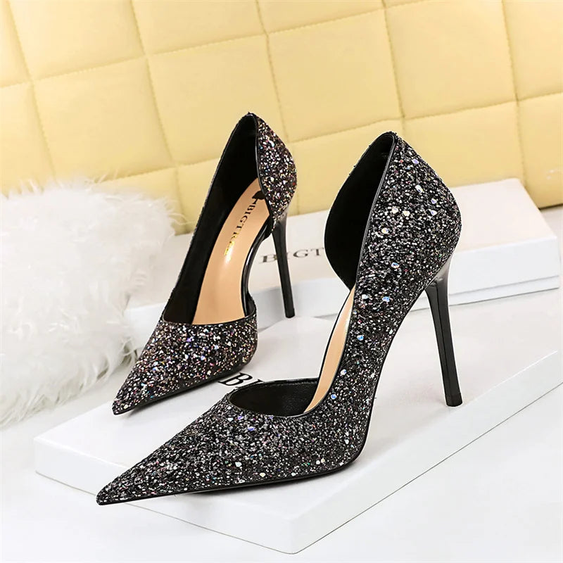 Black high-heeled pumps covered in silver glitter, displayed on a white surface against a soft, beige quilted backdrop, exuding elegance and glamor.