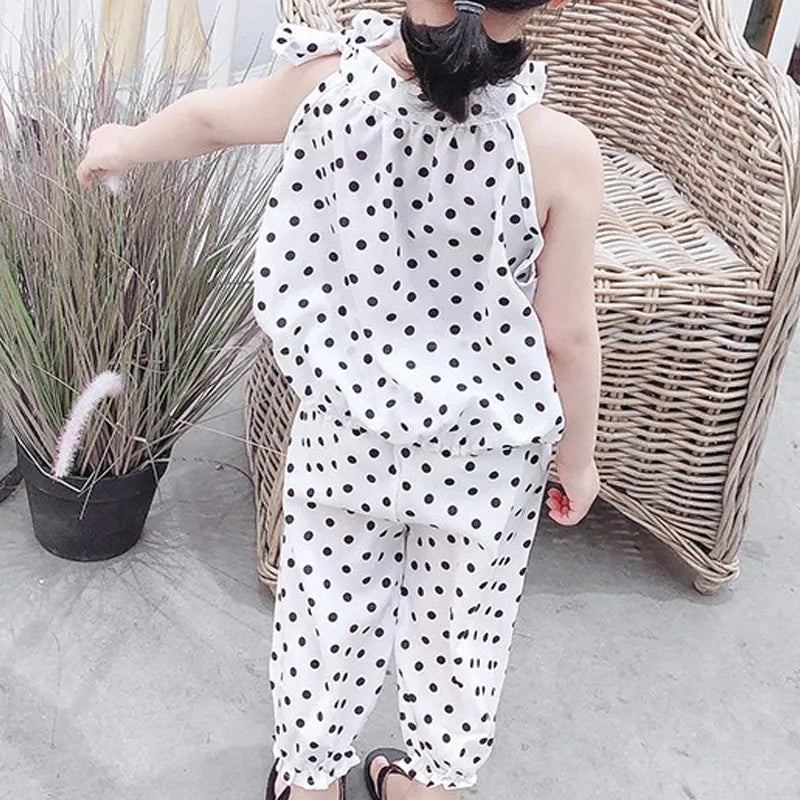 A child in a white polka dot outfit stands facing away, touching tall grasses. A wicker chair is beside them on a concrete surface.