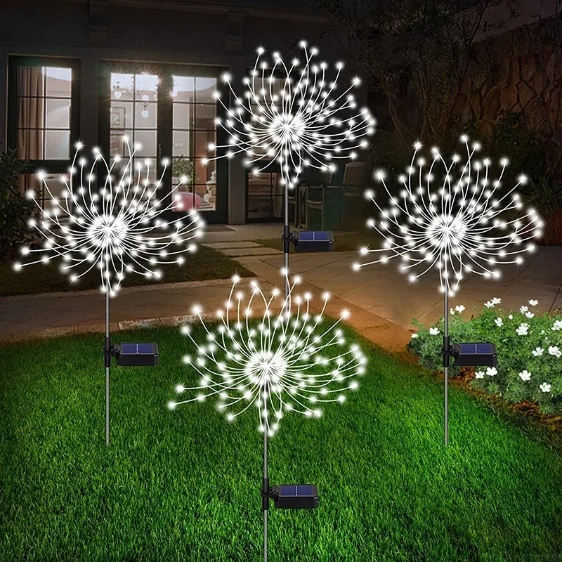 Solar-powered LED lights resembling dandelions illuminate a grassy yard at night. The house in the background features lit windows, and flowers are visible on the right.