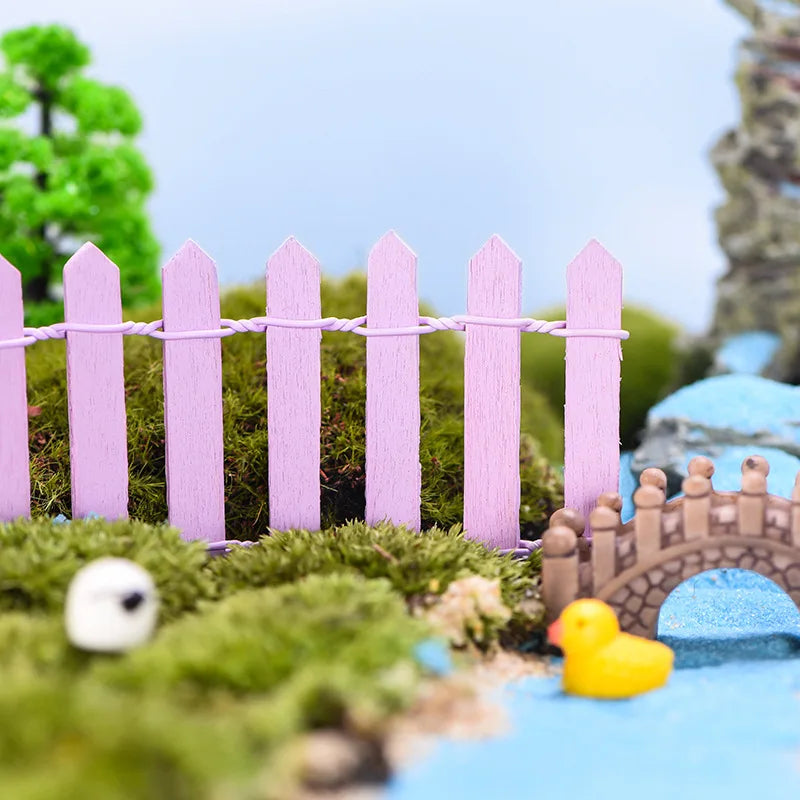 Miniature scene with a pink picket fence, green moss, a small bridge, and a rubber duck near a bright blue pond. A tree and rocks complete the diorama under a clear sky.