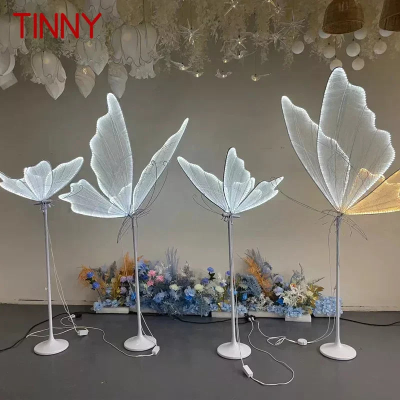 Four decorative floor lamps resembling delicate, translucent leaves emit a soft glow. They are arranged on a dark floor with floral arrangements and hanging ornaments above.